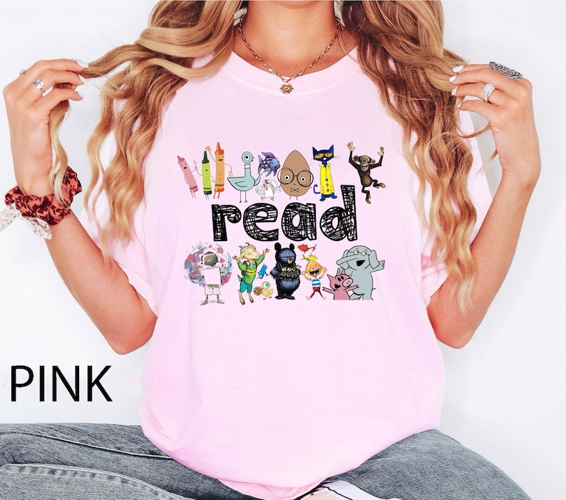 May include: A light pink t-shirt featuring the word read in a black scribbled font. The design includes colorful illustrations of various children's book characters, including a pigeon, a cat, a monkey, an elephant, and a pig, arranged above and below the text.
