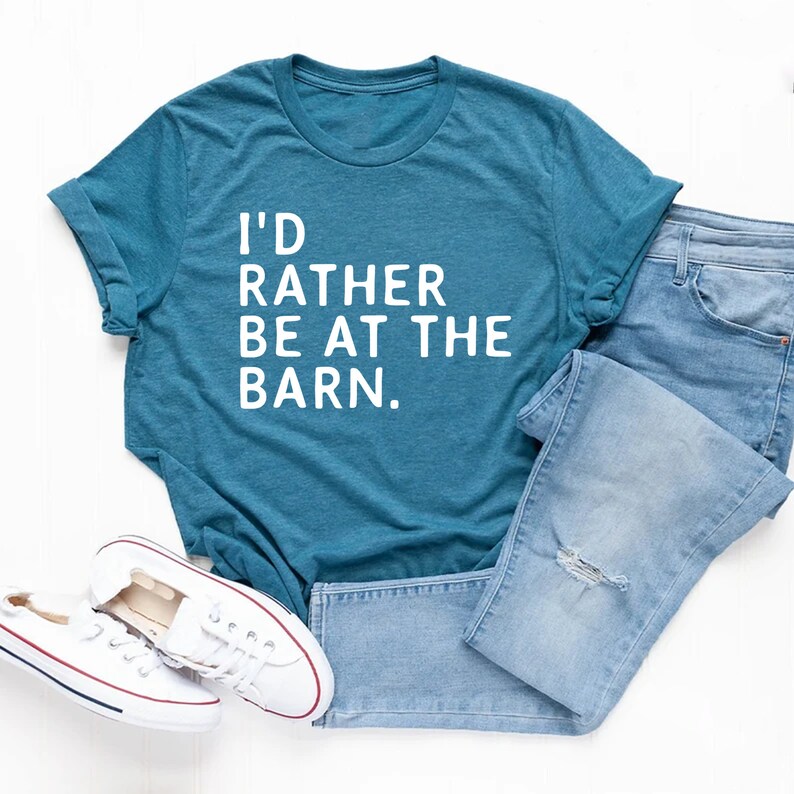 Puede incluir: Camiseta de color turquesa con el texto "I'D RATHER BE AT THE BARN." Se combina con vaqueros azules claros y zapatillas de lona blancas. La camiseta tiene las mangas enrolladas.