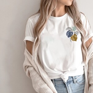 May include: White t-shirt featuring a minimalist botanical design of stylized leaves in shades of blue and brown. The shirt is made of a soft material and is suitable for casual wear. The sleeves are rolled up, and the shirt is knotted at the waist.