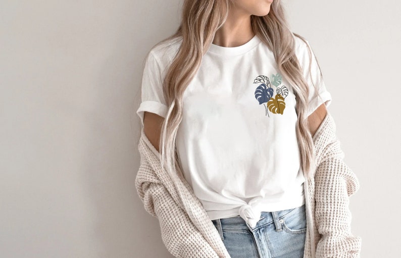 May include: White t-shirt featuring a minimalist botanical design of stylized leaves in shades of blue and brown. The shirt is made of a soft material and is suitable for casual wear. The sleeves are rolled up, and the shirt is knotted at the waist.