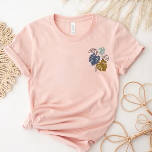 May include: A light pink t-shirt with rolled-up sleeves. The shirt features a small, minimalist design of tropical leaves in shades of blue, green, and brown. The shirt is made of soft material and is suitable for casual wear.