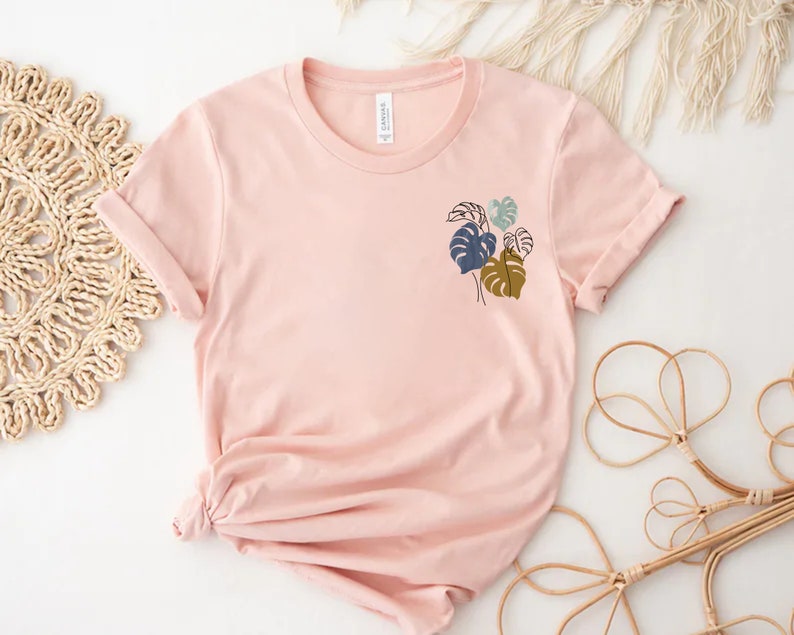 May include: A light pink t-shirt with rolled-up sleeves. The shirt features a small, minimalist design of tropical leaves in shades of blue, green, and brown. The shirt is made of soft material and is suitable for casual wear.