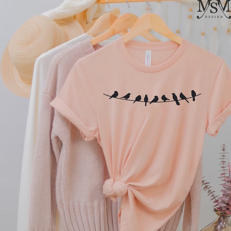May include: A peach-colored t-shirt with a black silhouette of a line of birds on a wire. The shirt is hanging on a wooden hanger.