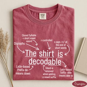 May include: A crimson short-sleeved t-shirt featuring white text that reads The shirt is decodable, surrounded by white arrows and labels explaining linguistic concepts like Closed Syllable, Digraphs, r-controlled, and Latin-based suffixes.