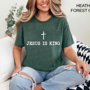 K&ouml;nnte beinhalten: Ein dunkelgr&uuml;nes T-Shirt mit dem wei&szlig;en Text "JESUS IS KING" und einem wei&szlig;en Kreuz. Das T-Shirt hat einen Rundhalsausschnitt.