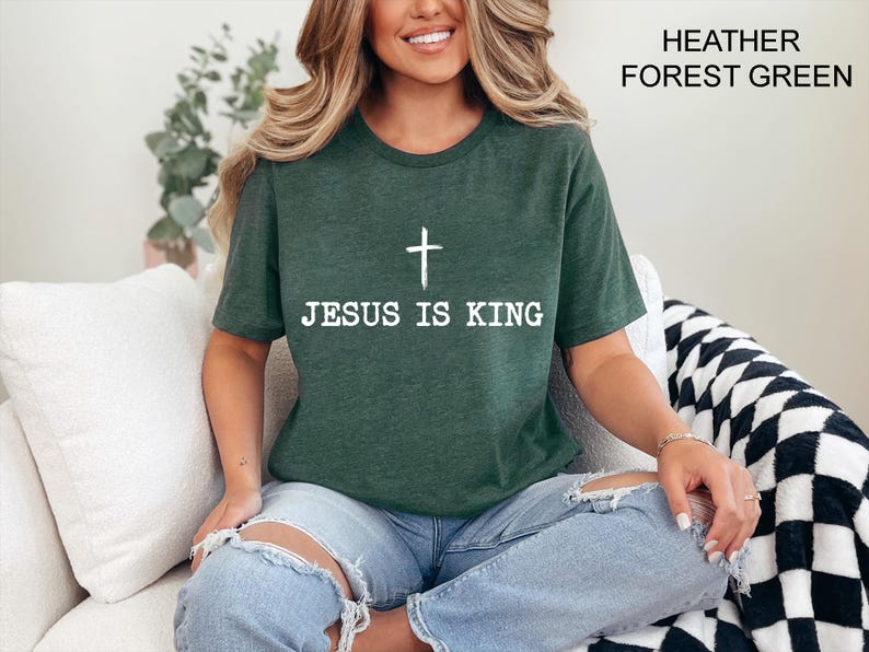 K&ouml;nnte beinhalten: Ein dunkelgr&uuml;nes T-Shirt mit dem wei&szlig;en Text "JESUS IS KING" und einem wei&szlig;en Kreuz. Das T-Shirt hat einen Rundhalsausschnitt.
