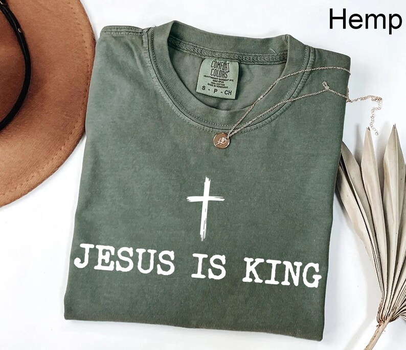 K&ouml;nnte beinhalten: Olivgr&uuml;nes T-Shirt mit dem wei&szlig;en Text &bdquo;JESUS IS KING&ldquo; und einem wei&szlig;en Kreuz. Eine goldene Halskette mit Anh&auml;nger liegt auf dem Shirt. Das Wort &bdquo;Hemp&ldquo; befindet sich in der oberen rechten Ecke.