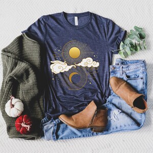 May include: A navy blue t-shirt featuring a gold sun and moon design. The design includes clouds and stars.