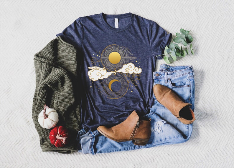 May include: A navy blue t-shirt featuring a gold sun and moon design. The design includes clouds and stars.