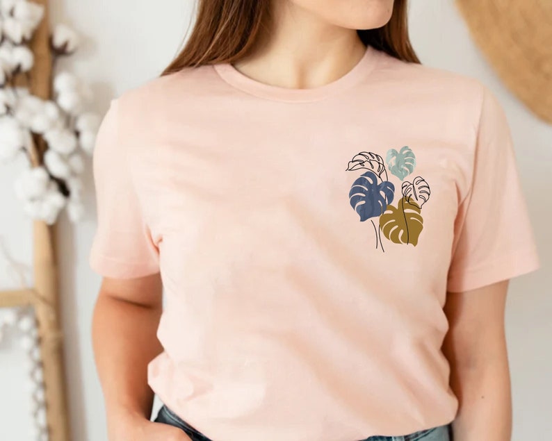 May include: A light pink t-shirt featuring a minimalist botanical design. The design includes stylized leaves in shades of navy blue, olive green, and light blue. The shirt has short sleeves and a crew neck.