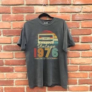 May include: A charcoal grey short-sleeve t-shirt featuring a graphic print of a cassette tape above a sunset design. The text reads "Vintage 1976 LIMITED EDITION" in a retro font with orange, yellow, and teal stripes.