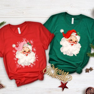 May include: Two Christmas-themed t-shirts, one red and one green, each featuring a vintage Santa Claus graphic. The Santa has a white beard, rosy cheeks, and a red hat. The red shirt has snowflakes around the Santa face.