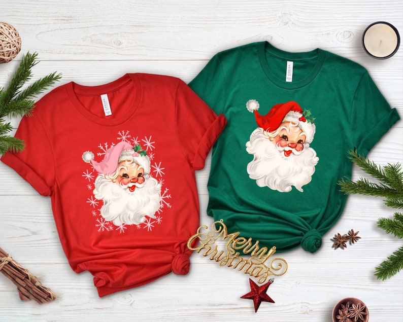 May include: Two Christmas-themed t-shirts, one red and one green, each featuring a vintage Santa Claus graphic. The Santa has a white beard, rosy cheeks, and a red hat. The red shirt has snowflakes around the Santa face.