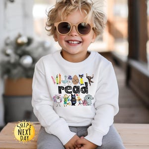 May include: A white long-sleeve sweatshirt featuring a graphic print of various colorful cartoon characters and the word read in a black patterned font. The design includes illustrations of a crayon, a bird, a cat, a monkey, and other figures.
