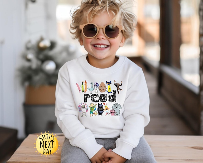 May include: A white long-sleeve sweatshirt featuring a graphic print of various colorful cartoon characters and the word read in a black patterned font. The design includes illustrations of a crayon, a bird, a cat, a monkey, and other figures.