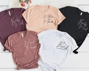 May include: A collection of five short-sleeved crew neck t-shirts in maroon, peach, black, mauve, and white. Each shirt features cursive text on the left chest area reading Bride, Bridesmaid, Maid of Honor, or Matron of Honor in black or white ink.