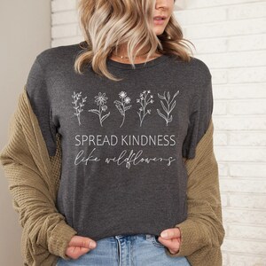 Op de afbeelding: Een donkergrijs T-shirt met een witte afbeelding van wilde bloemen en de tekst "SPREAD KINDNESS like wildflowers".