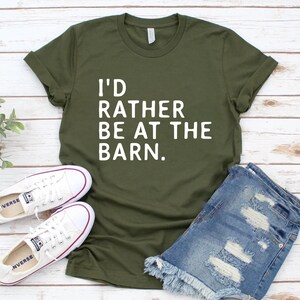 Puede incluir: Camiseta verde oliva con el texto "I'D RATHER BE AT THE BARN." La imagen tambi&eacute;n muestra pantalones cortos de mezclilla y zapatillas de lona blancas. El atuendo informal se muestra sobre una superficie de madera clara.