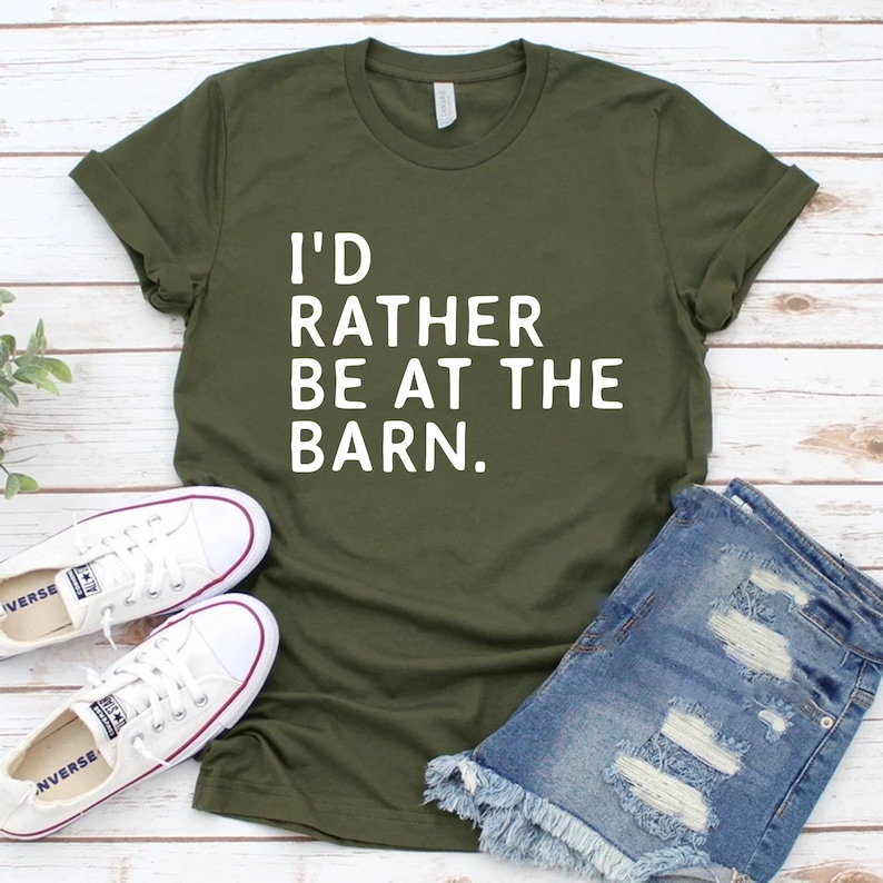 Puede incluir: Camiseta verde oliva con el texto "I'D RATHER BE AT THE BARN." La imagen tambi&eacute;n muestra pantalones cortos de mezclilla y zapatillas de lona blancas. El atuendo informal se muestra sobre una superficie de madera clara.