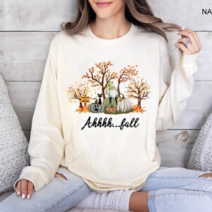 May include: A natural-colored long-sleeve sweatshirt featuring a graphic print of autumn trees, pumpkins, a black cat, and a gnome. The text "Ahhhh...fall" is printed in black script below the illustration. The word "NATURAL" appears in the top corner.