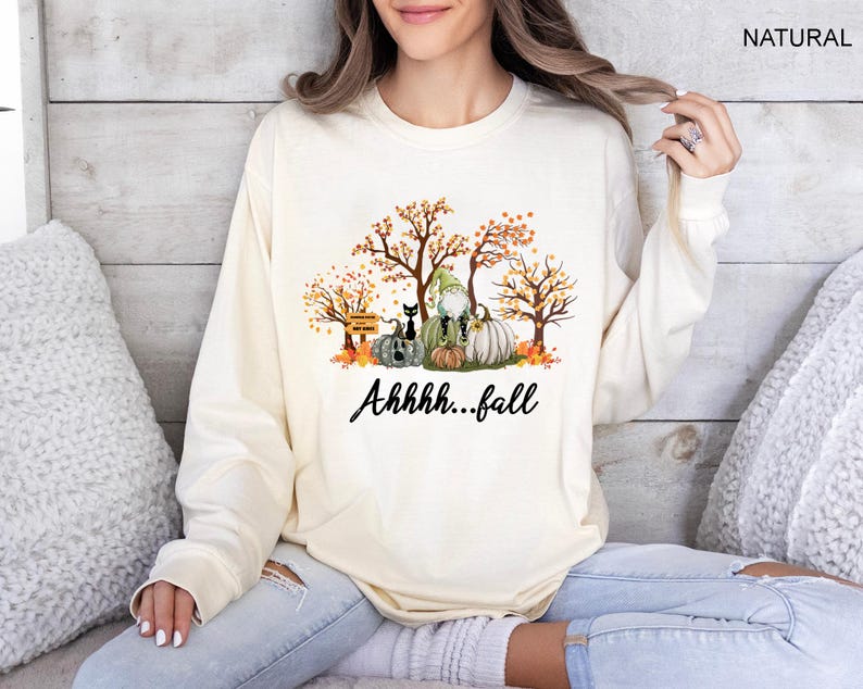 May include: A natural-colored long-sleeve sweatshirt featuring a graphic print of autumn trees, pumpkins, a black cat, and a gnome. The text "Ahhhh...fall" is printed in black script below the illustration. The word "NATURAL" appears in the top corner.
