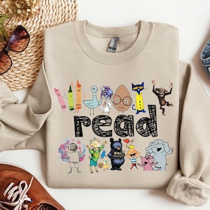 May include: A beige crewneck sweatshirt featuring a graphic print of various colorful cartoon characters and the word "read" in a black, hand-drawn style font. The design includes illustrations of crayons, a pigeon, a cat, a monkey, and other figures.