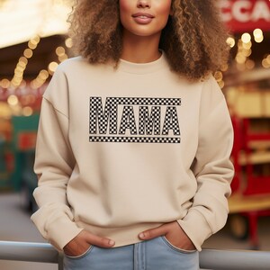Puede incluir: Sudadera beige con la palabra "MAMA" en un dise&ntilde;o a cuadros blanco y negro. La sudadera tiene mangas largas y cuello redondo. El texto est&aacute; centrado en el pecho. La modelo lleva vaqueros azules.