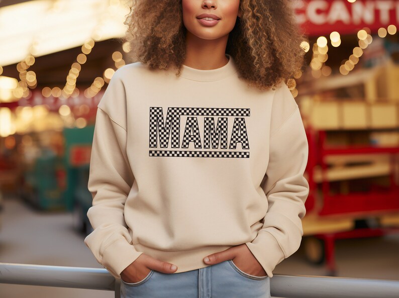 Puede incluir: Sudadera beige con la palabra "MAMA" en un dise&ntilde;o a cuadros blanco y negro. La sudadera tiene mangas largas y cuello redondo. El texto est&aacute; centrado en el pecho. La modelo lleva vaqueros azules.