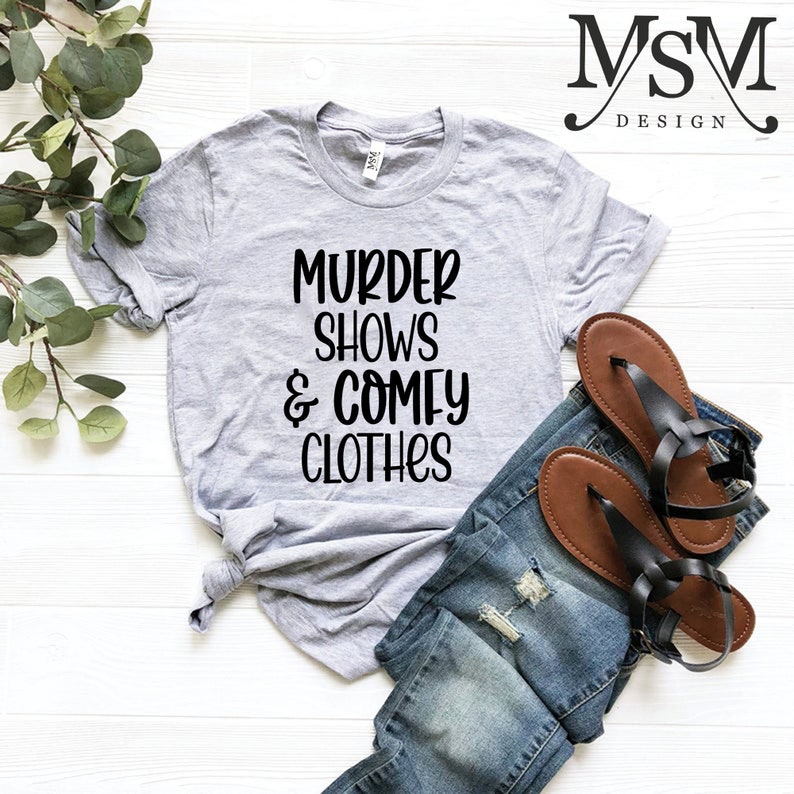 May include: A heather gray t-shirt with the text "MURDER SHOWS & COMFY CLOTHES" in black. The shirt is paired with blue denim jeans and brown sandals with black straps. The image is styled on a white surface with green foliage.