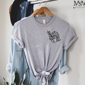Peut inclure: Un t-shirt gris chin&eacute; orn&eacute; d'un motif noir de deux champignons &agrave; chapeaux tachet&eacute;s et de feuilles. Le t-shirt est suspendu &agrave; un cintre en bois, avec une chemise en jean et un chapeau en arri&egrave;re-plan.