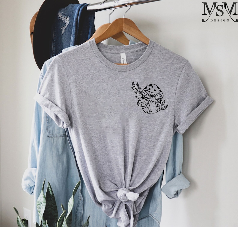 Peut inclure: Un t-shirt gris chin&eacute; orn&eacute; d'un motif noir de deux champignons &agrave; chapeaux tachet&eacute;s et de feuilles. Le t-shirt est suspendu &agrave; un cintre en bois, avec une chemise en jean et un chapeau en arri&egrave;re-plan.
