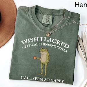 May include: A sage green crew neck t-shirt featuring a graphic of a standing frog holding a small orange mug. The white text reads: WISH I LACKED CRITICAL THINKING SKILLS Y'ALL SEEM SO HAPPY. The shirt has a Comfort Colors brand label at the neck.