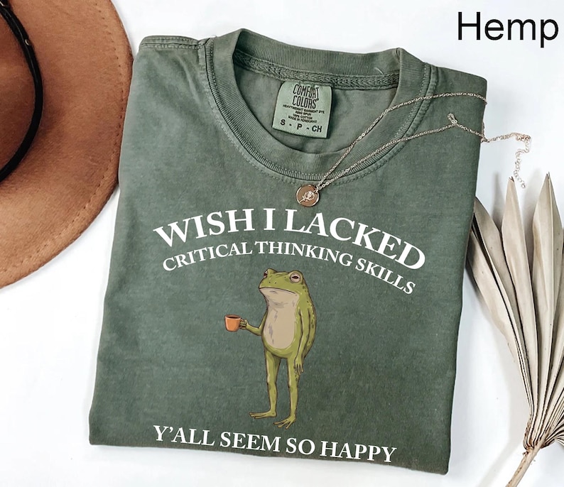 May include: A sage green crew neck t-shirt featuring a graphic of a standing frog holding a small orange mug. The white text reads: WISH I LACKED CRITICAL THINKING SKILLS Y'ALL SEEM SO HAPPY. The shirt has a Comfort Colors brand label at the neck.