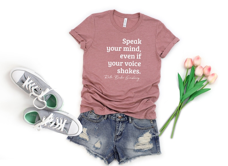 May include: A pink t-shirt with the quote 'Speak your mind, even if your voice shakes. Ruth Bader Ginsburg' printed in white. The t-shirt is paired with blue denim shorts and gray sneakers. A bouquet of pink tulips with green stems is in the background.