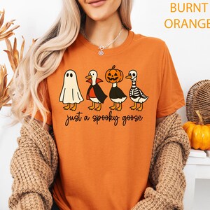 Puede incluir: Camiseta naranja quemado con un dise&ntilde;o de Halloween. El gr&aacute;fico muestra cuatro gansos de dibujos animados disfrazados de fantasma, vampiro, calabaza y esqueleto. El texto "just a spooky goose" est&aacute; impreso debajo.