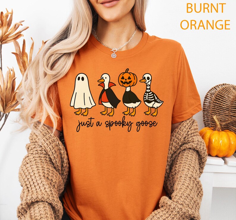 Puede incluir: Camiseta naranja quemado con un dise&ntilde;o de Halloween. El gr&aacute;fico muestra cuatro gansos de dibujos animados disfrazados de fantasma, vampiro, calabaza y esqueleto. El texto "just a spooky goose" est&aacute; impreso debajo.