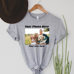 Może przedstawiać: Szara koszulka z nadrukowanym zdjęciem. Zdjęcie przedstawia rodzinę z psem, a tekst "Your Photo Here" i "Your text here" jest wydrukowany nad i pod zdjęciem. Koszulka wisi na drewnianym wieszaku.