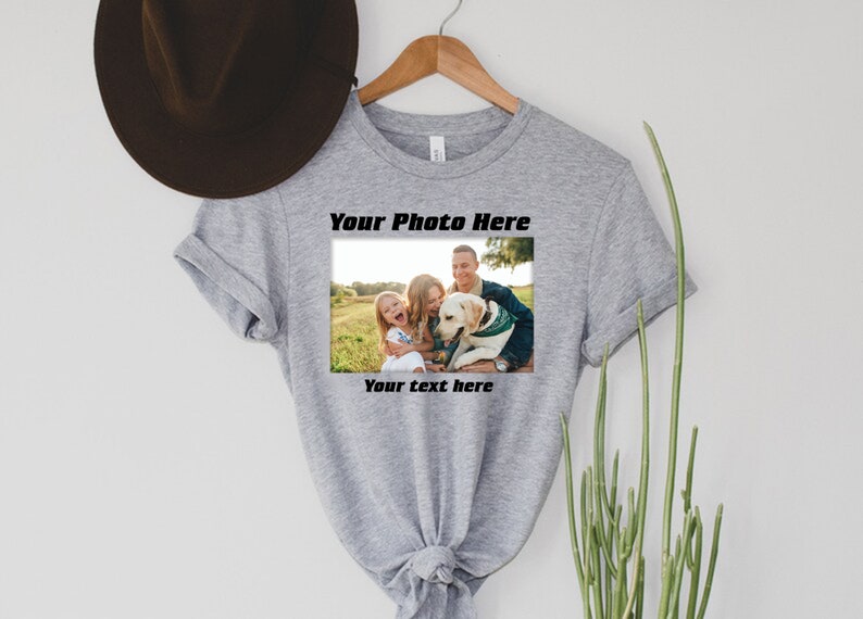 Może przedstawiać: Szara koszulka z nadrukowanym zdjęciem. Zdjęcie przedstawia rodzinę z psem, a tekst "Your Photo Here" i "Your text here" jest wydrukowany nad i pod zdjęciem. Koszulka wisi na drewnianym wieszaku.