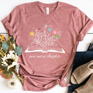 May include: A pink t-shirt with a white graphic of an open book with flowers growing out of it. The text "one more chapter" is printed below the book.