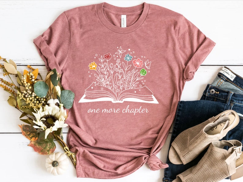 May include: A pink t-shirt with a white graphic of an open book with flowers growing out of it. The text "one more chapter" is printed below the book.