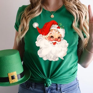 May include: Green t-shirt featuring a vintage Santa Claus graphic with a red hat and white beard. A green top hat with a gold buckle is held in the image. The shirt is a festive holiday apparel item.