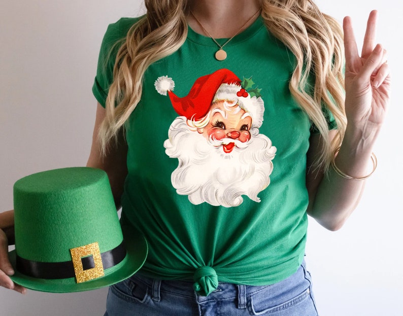 May include: Green t-shirt featuring a vintage Santa Claus graphic with a red hat and white beard. A green top hat with a gold buckle is held in the image. The shirt is a festive holiday apparel item.