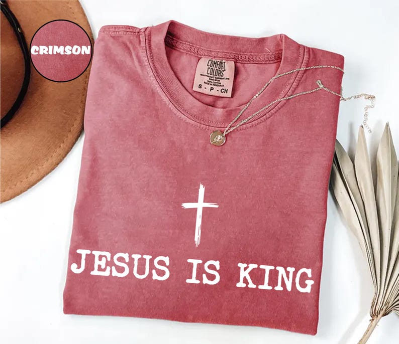K&ouml;nnte beinhalten: Karmesinrotes T-Shirt mit dem Text "JESUS IS KING" und einer wei&szlig;en Kreuzgrafik. Das Shirt hat einen Rundhalsausschnitt und kurze &Auml;rmel. Eine goldene Halskette mit Anh&auml;nger liegt &uuml;ber dem Shirt. Das Shirt ist aus einem weichen Material.