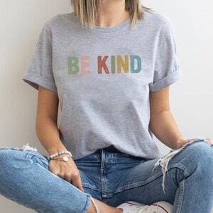 May include: A light gray t-shirt with the words "BE KIND" printed across the chest in pastel colors. The shirt has rolled-up sleeves and is paired with blue jeans. The model is sitting cross-legged.