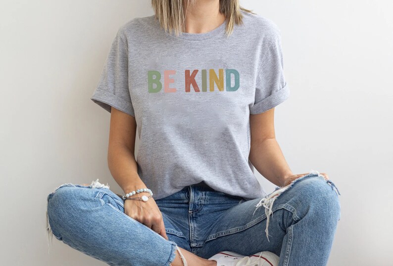 May include: A light gray t-shirt with the words "BE KIND" printed across the chest in pastel colors. The shirt has rolled-up sleeves and is paired with blue jeans. The model is sitting cross-legged.