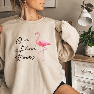 K&ouml;nnte beinhalten: Beiges Sweatshirt mit dem Text "Our Flock Rocks" und einer rosa Flamingo-Grafik. Der Flamingo steht auf einem Bein. Das Sweatshirt hat eine neutrale Farbe und einen Rundhalsausschnitt.