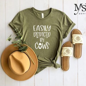 May include: Olive green t-shirt with the text "EASILY DISTRACTED BY COWS" in white. A brown hat and a pair of cream-colored sandals are next to the shirt. The shirt is knotted at the bottom. The background is a white wooden surface.