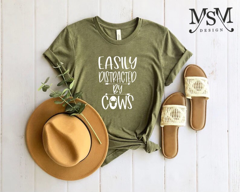 May include: Olive green t-shirt with the text "EASILY DISTRACTED BY COWS" in white. A brown hat and a pair of cream-colored sandals are next to the shirt. The shirt is knotted at the bottom. The background is a white wooden surface.