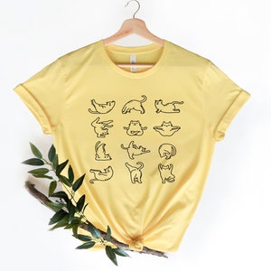 Op de afbeelding: Een geel T-shirt met een afbeelding van katten die yoga-houdingen aannemen. Het shirt heeft een ronde hals en korte mouwen.