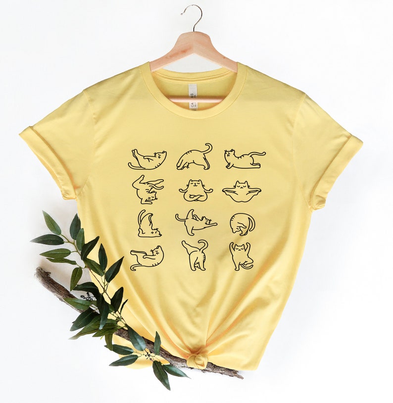 Op de afbeelding: Een geel T-shirt met een afbeelding van katten die yoga-houdingen aannemen. Het shirt heeft een ronde hals en korte mouwen.